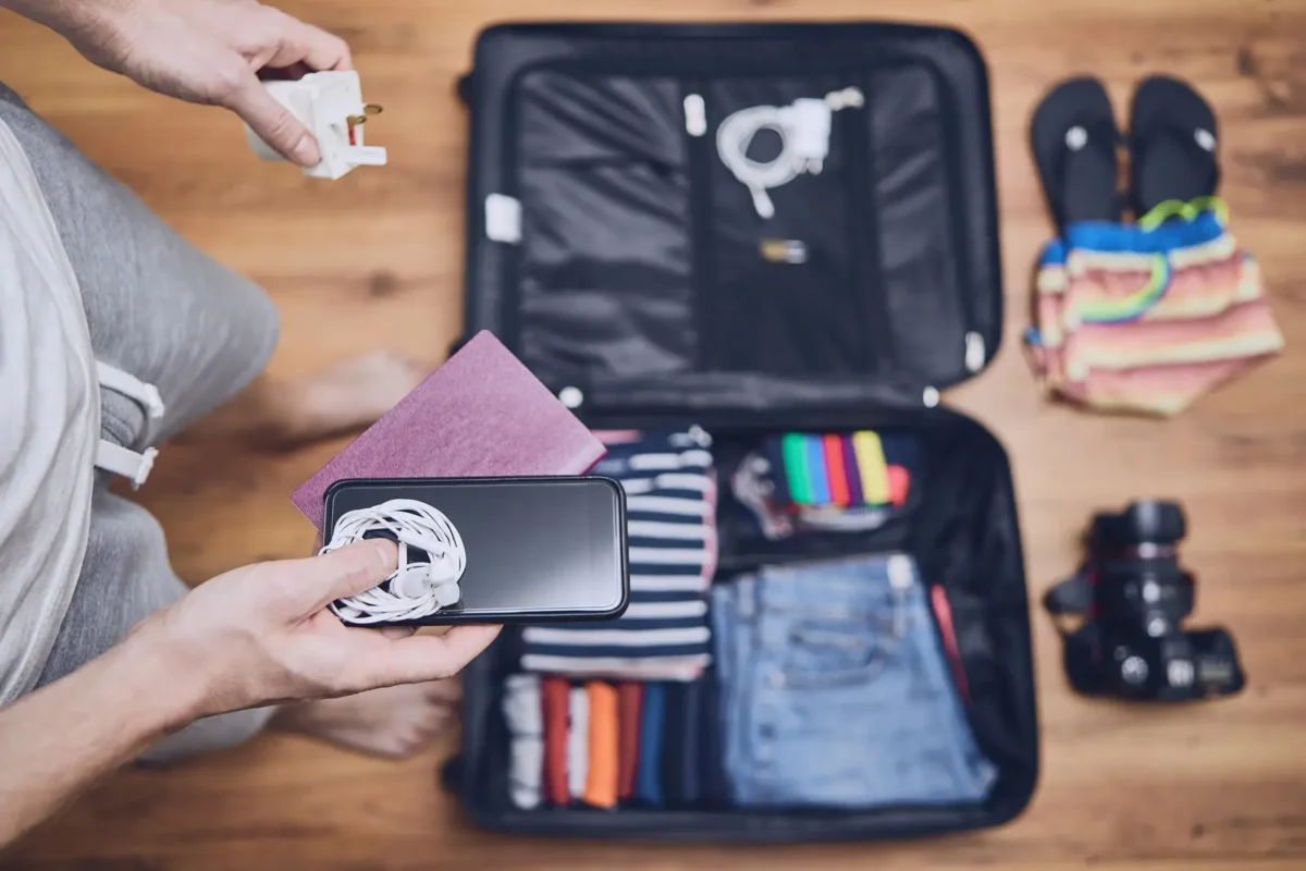 Personne préparant sa valise avec un passeport, un smartphone, un adaptateur et des accessoires de voyage, illustrant la planification d’un voyage.