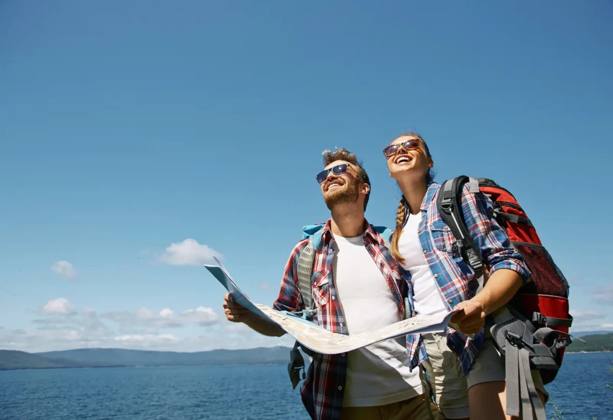Couple souriant avec sacs à dos et carte en main, explorant une destination sous un ciel bleu, symbolisant l'idée d'un voyage offert en cadeau.